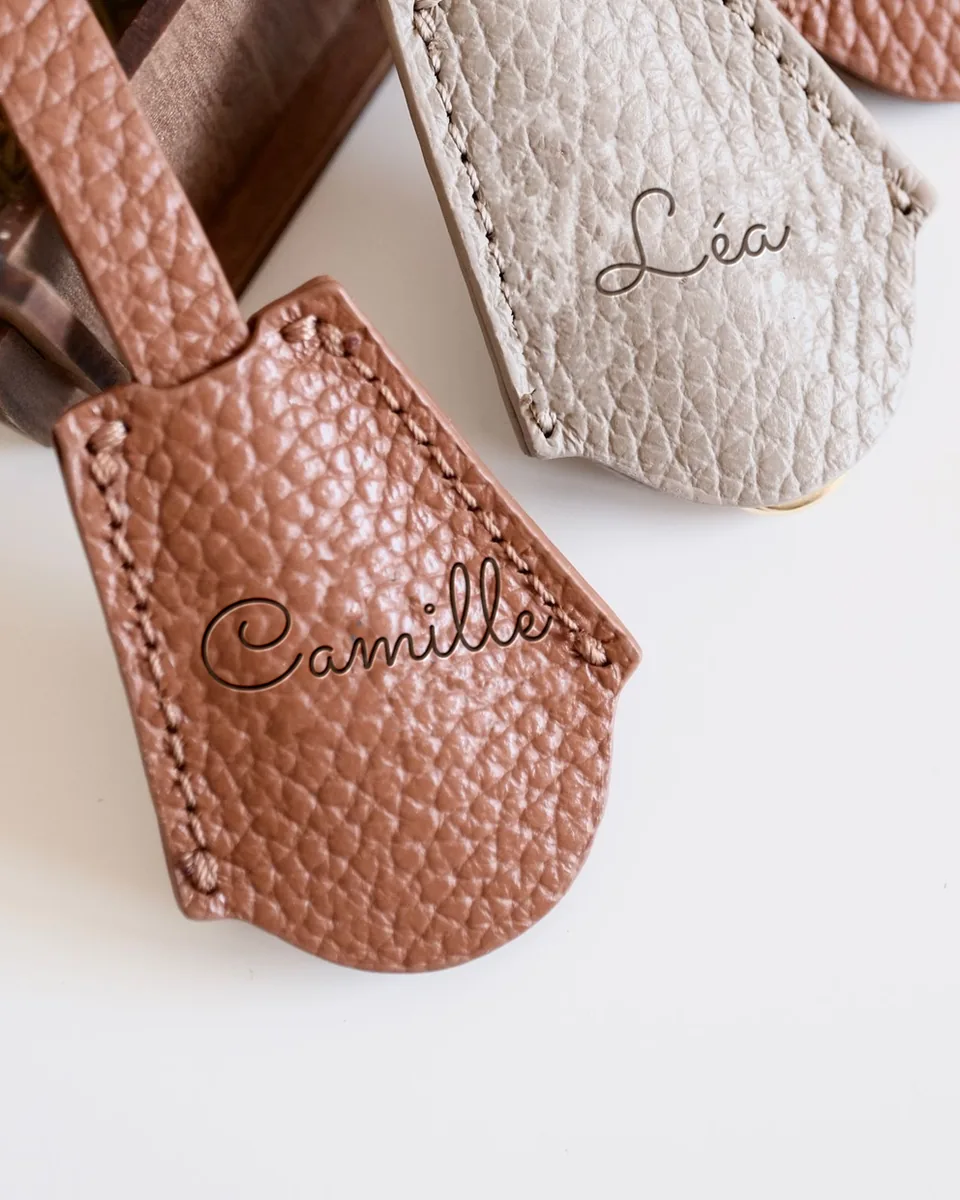 Breloques de sac en cuir grainé terracotta et beige, celle du premier plan gravée « Camille »