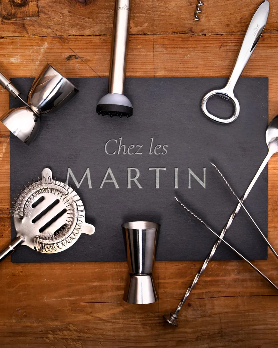 Outils de bar en inox autour d'un plateau d'ardoise gravé « Chez les MARTIN », sur une table en bois