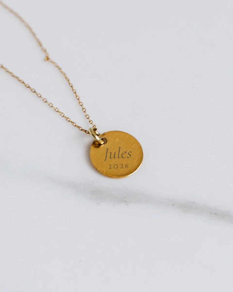 Médaille dorée gravée « Jules » et 2026, au bout d'une chaîne fine, sur du marbre