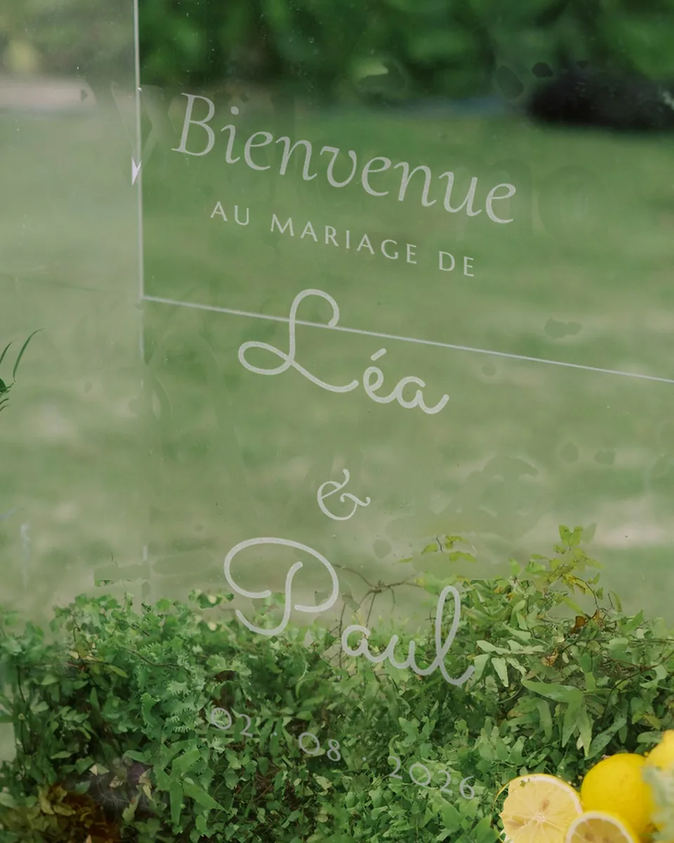 Panneau d'accueil en acrylique transparent gravé « Bienvenue au mariage de Léa & Paul », devant une haie et des citrons